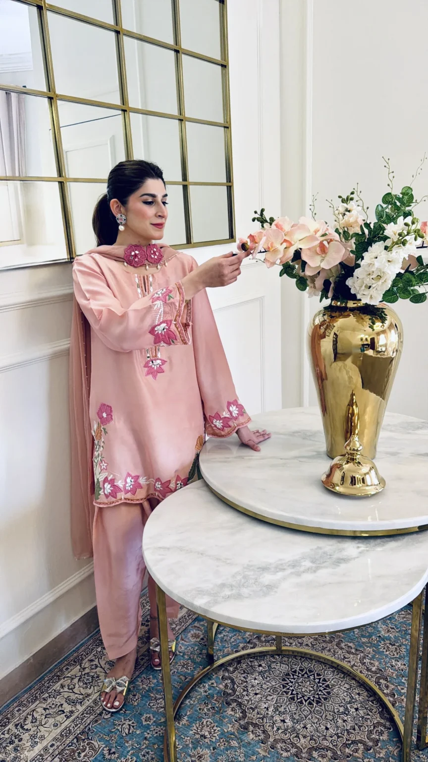 Three-quarter view of the salmon pink three-piece with fuchsia flowers on both lower sleeves and along the hem, a larger flower at the hip, and draped trousers