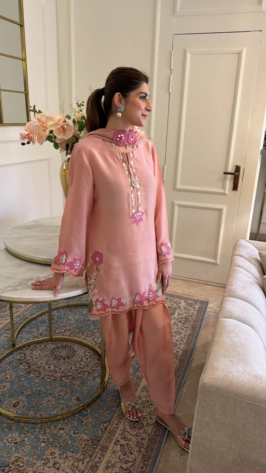 Short salmon pink shirt with a slit at the neck edged in tiny gold and pearl-toned trim, fuchsia flowers on the lower sleeves and along the hem, and a flower trail up one side, worn with draped trousers pointed at the ankle and a sheer matching dupatta