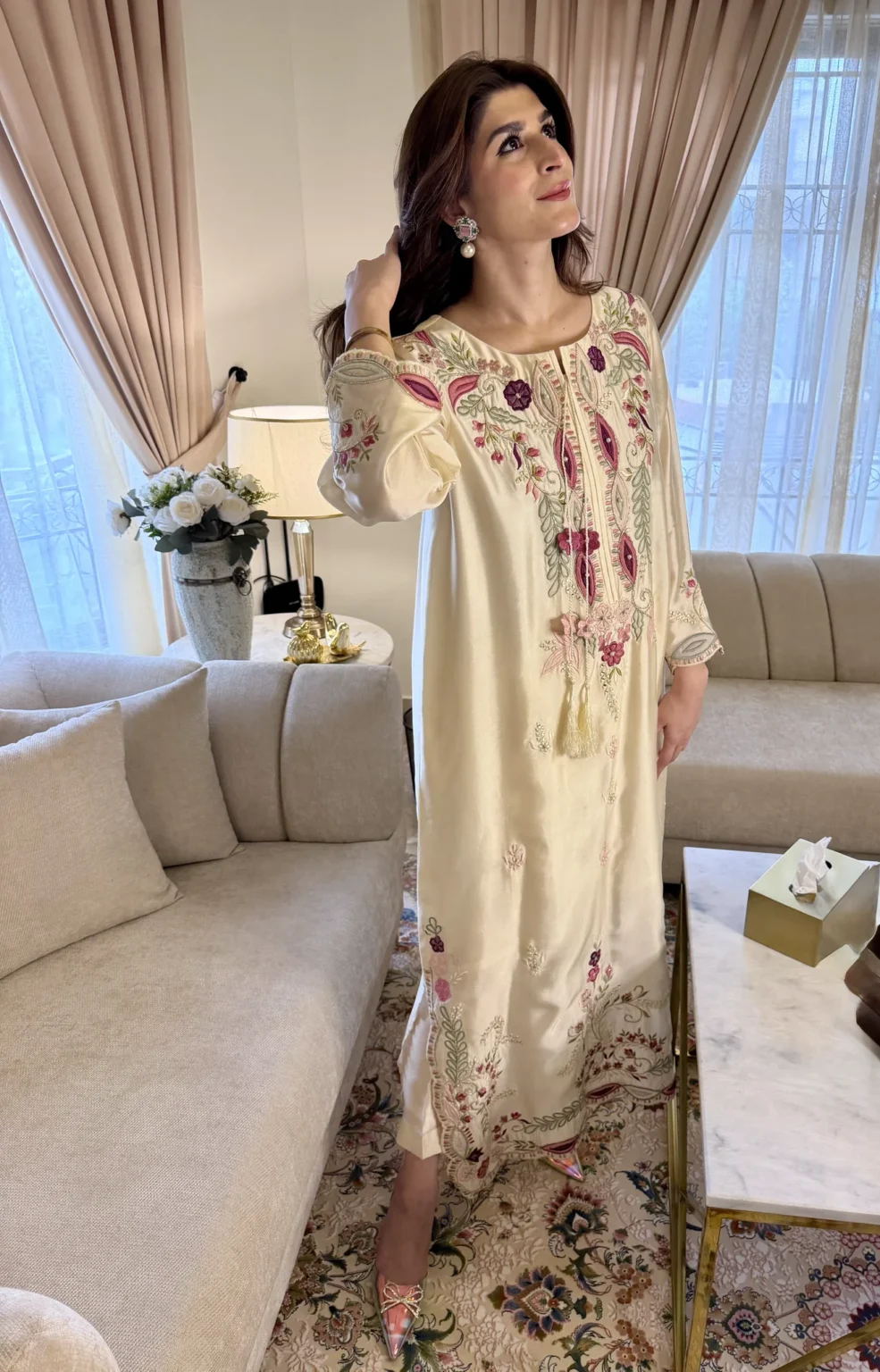 Angled view of the ivory silk kaftan with a spray of rose flowers and sage leaves above one cuff, tasseled cords, and the hem slit showing ivory trousers that stop above the ankle