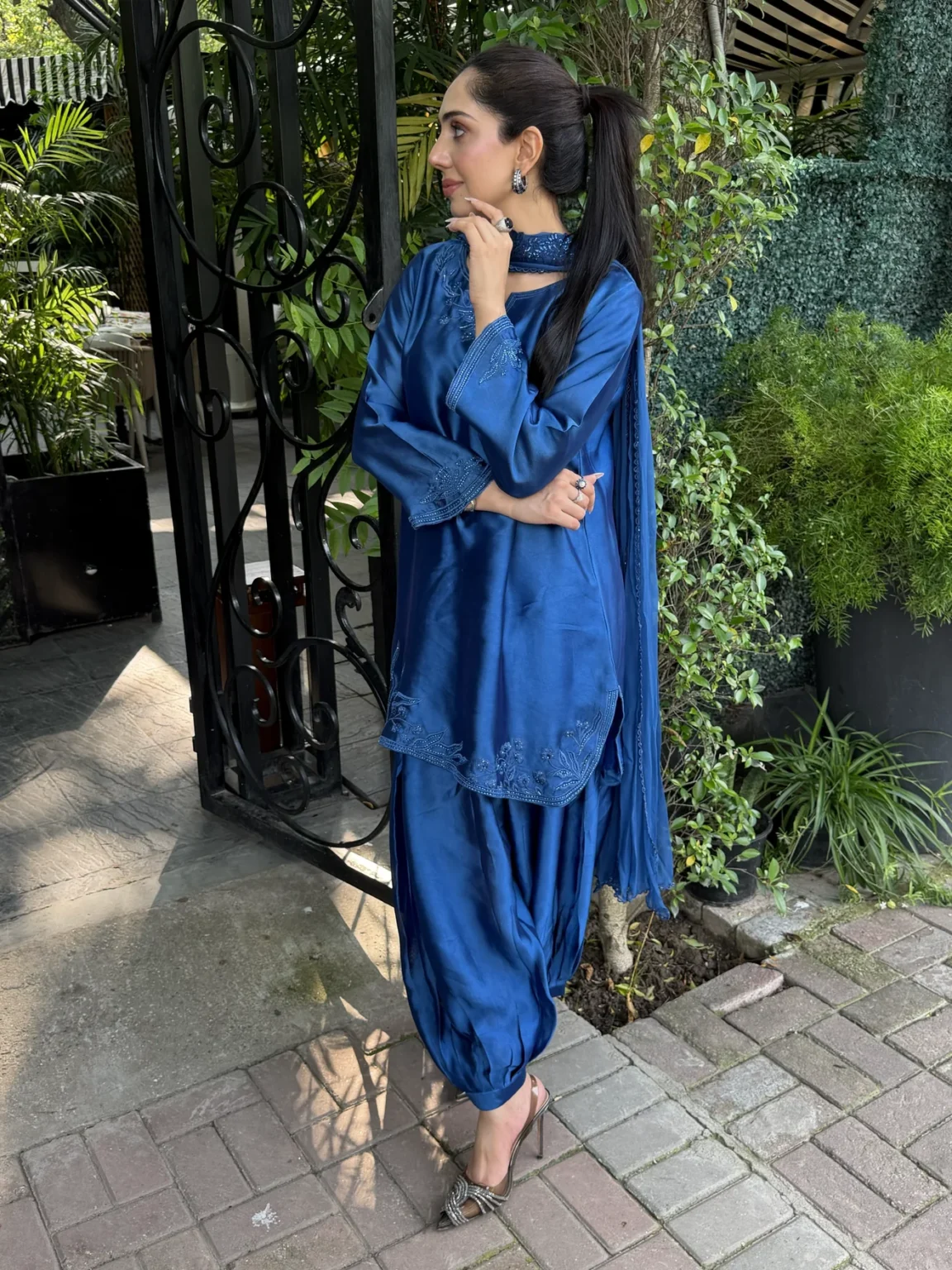 Three-quarter view of the sapphire blue silk shirt showing both embroidered cuffs with fine parallel rows of stitching and the side slit at the hem, with the dupatta's dotted trim beside the draped shalwar