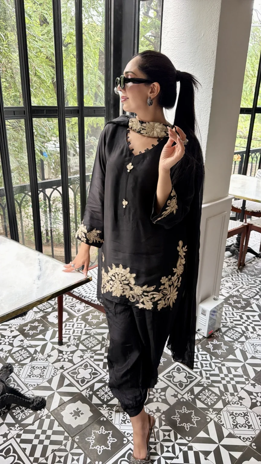 Short black silk shirt with a choker band of champagne embroidery at the throat, a keyhole neckline, embroidered cuffs and a floral border at the hem, with a loose black shalwar caught in at the ankle and a sheer black dupatta